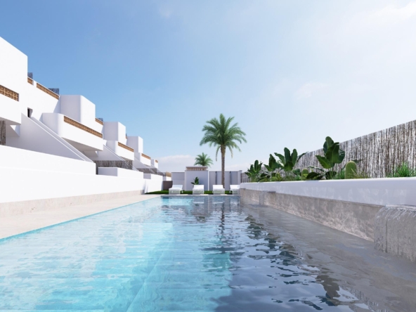 Modern white resort buildings line a rectangular pool with lounge chairs, under a clear blue sky.