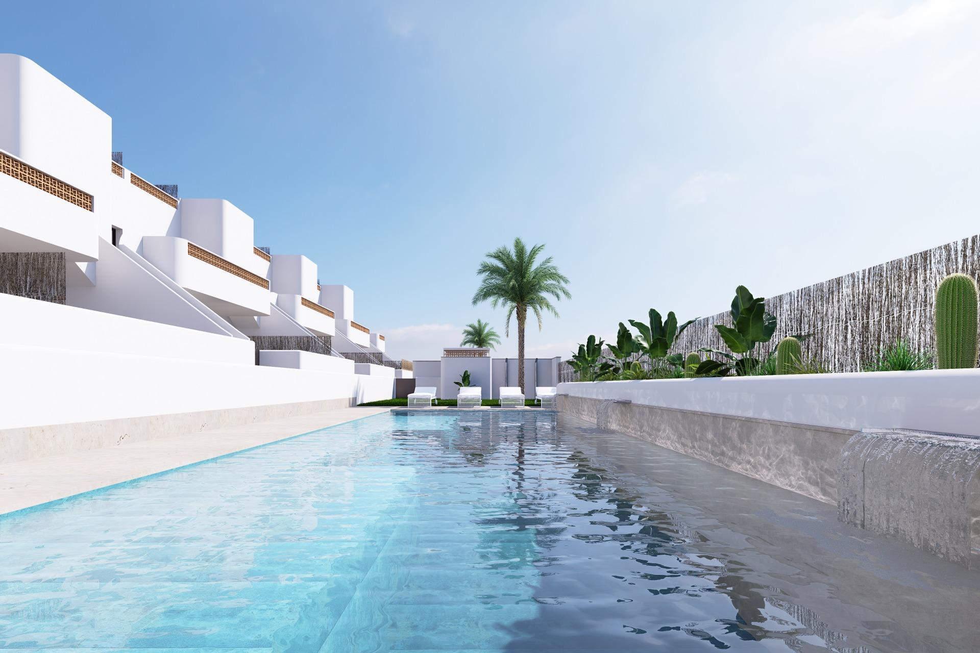 Modern white resort buildings line a rectangular pool with lounge chairs, under a clear blue sky.
