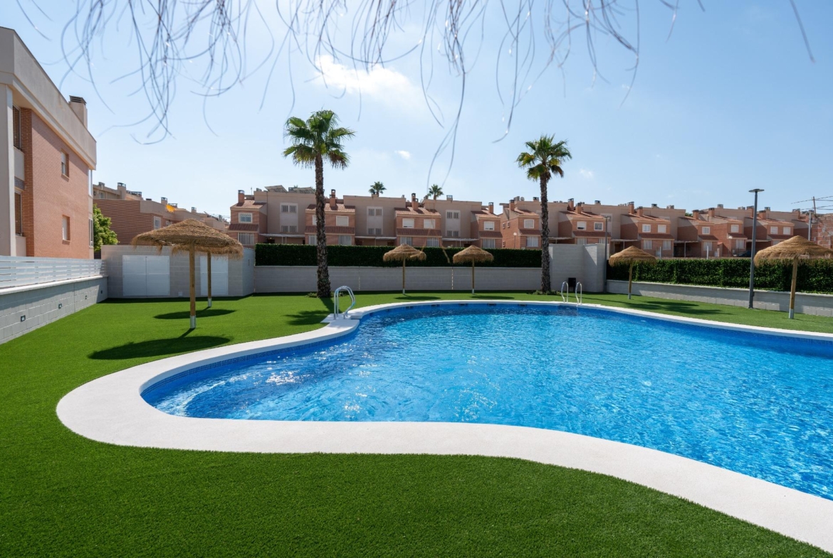 Rectangular blue swimming pool edged by white concrete, surrounded by green artificial grass, palm trees, and thatched shade umbrellas in a residential complex