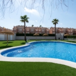 Rectangular blue swimming pool edged by white concrete, surrounded by green artificial grass, palm trees, and thatched shade umbrellas in a residential complex