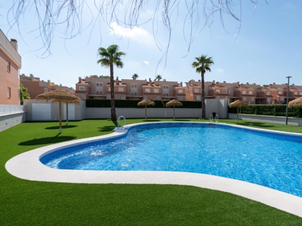 Rectangular blue swimming pool edged by white concrete, surrounded by green artificial grass, palm trees, and thatched shade umbrellas in a residential complex