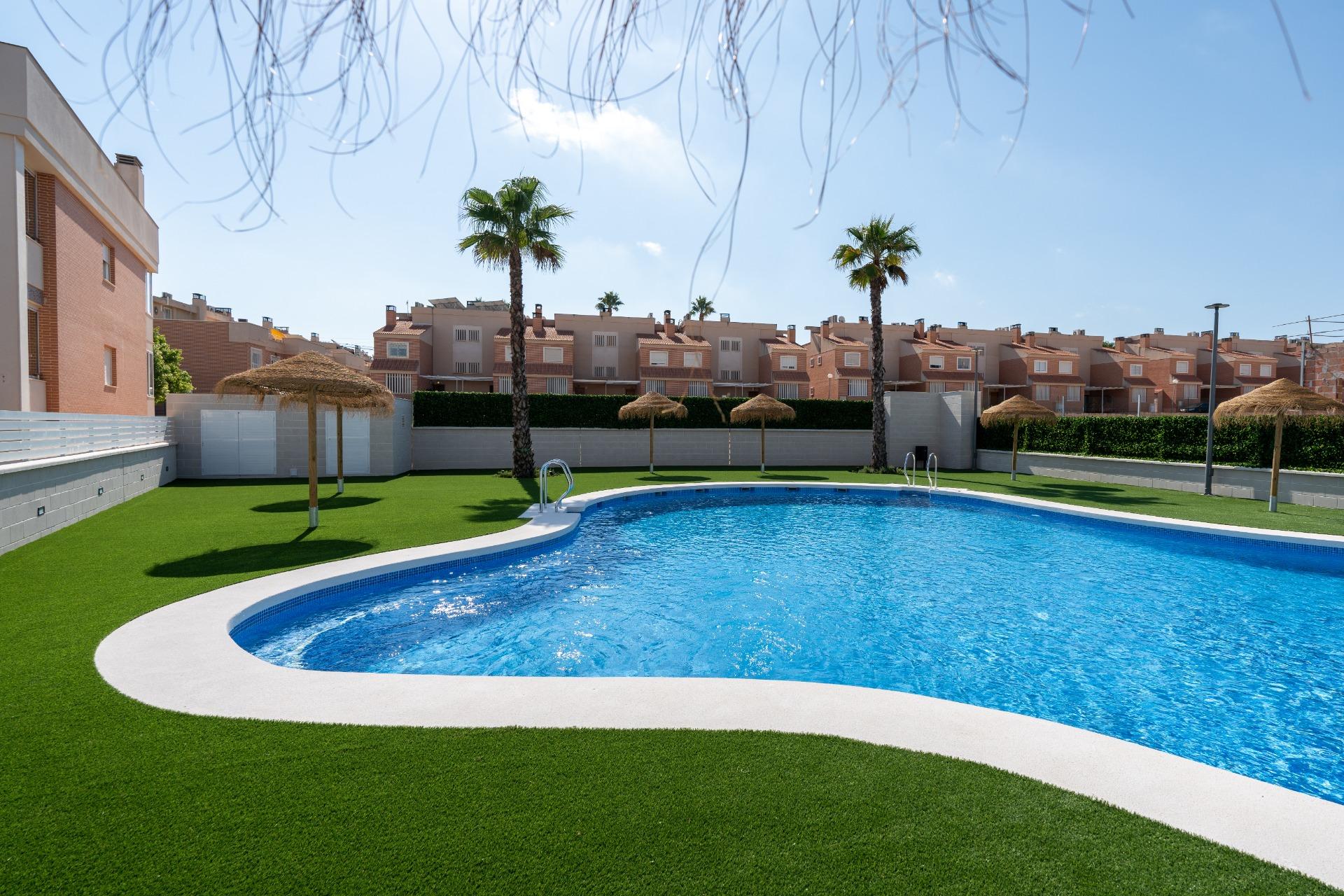 Rectangular blue swimming pool edged by white concrete, surrounded by green artificial grass, palm trees, and thatched shade umbrellas in a residential complex