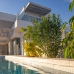 Modern white house with glass balconies by a rectangular pool and lush green trees under a clear blue sky.