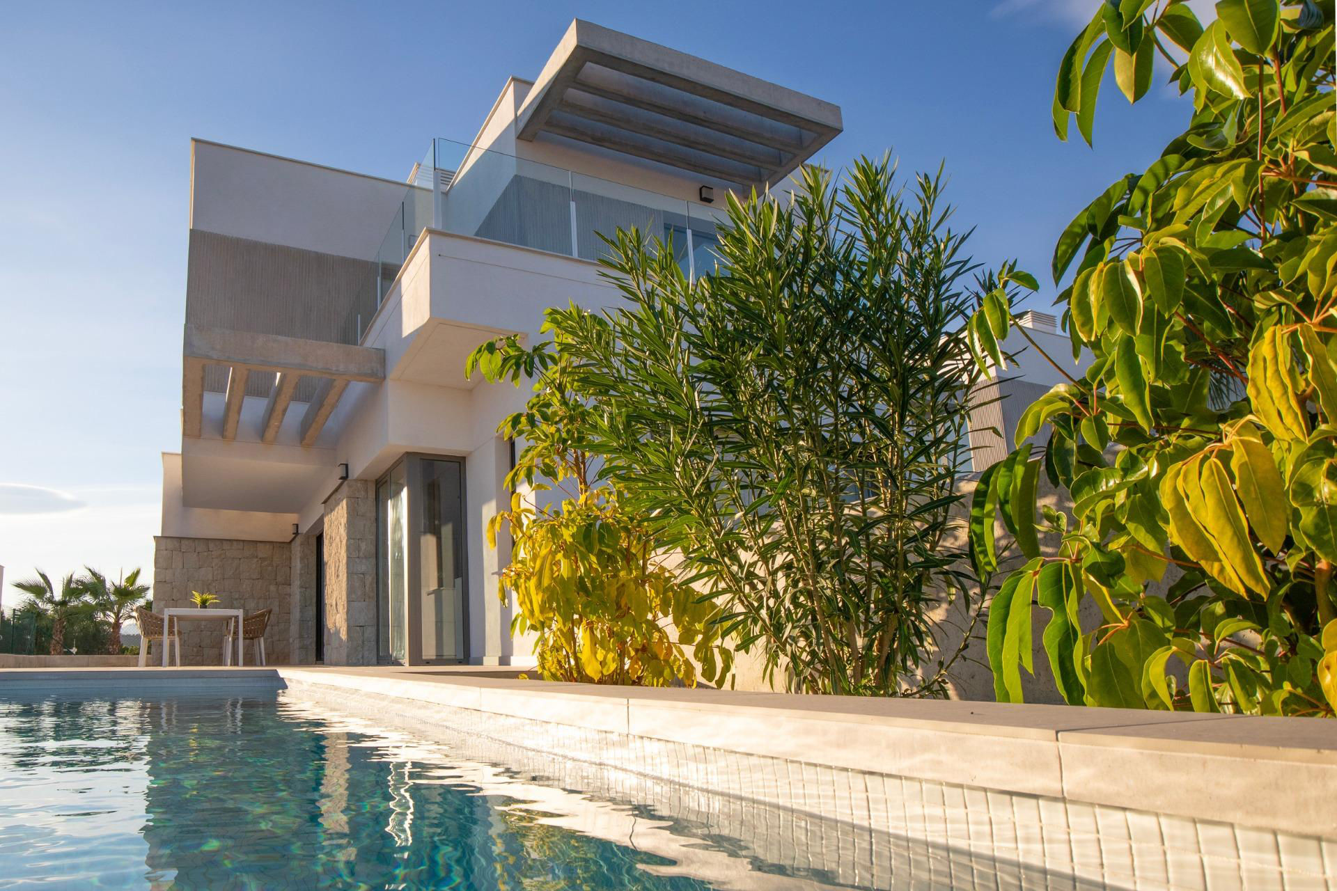 Modern white house with glass balconies by a rectangular pool and lush green trees under a clear blue sky.