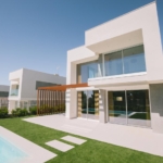 Modern white two-story villa with glass doors, a wooden pergola, and a rectangular pool in a sunny yard.