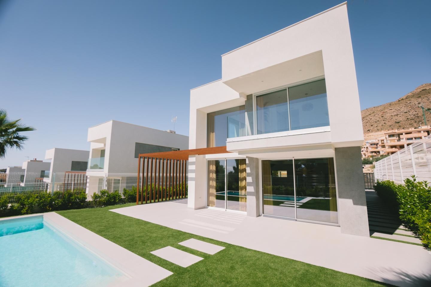 Modern white two-story villa with glass doors, a wooden pergola, and a rectangular pool in a sunny yard.