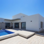 Modern white villa with a rectangular blue pool, glass doors, and a sunlit tiled patio.