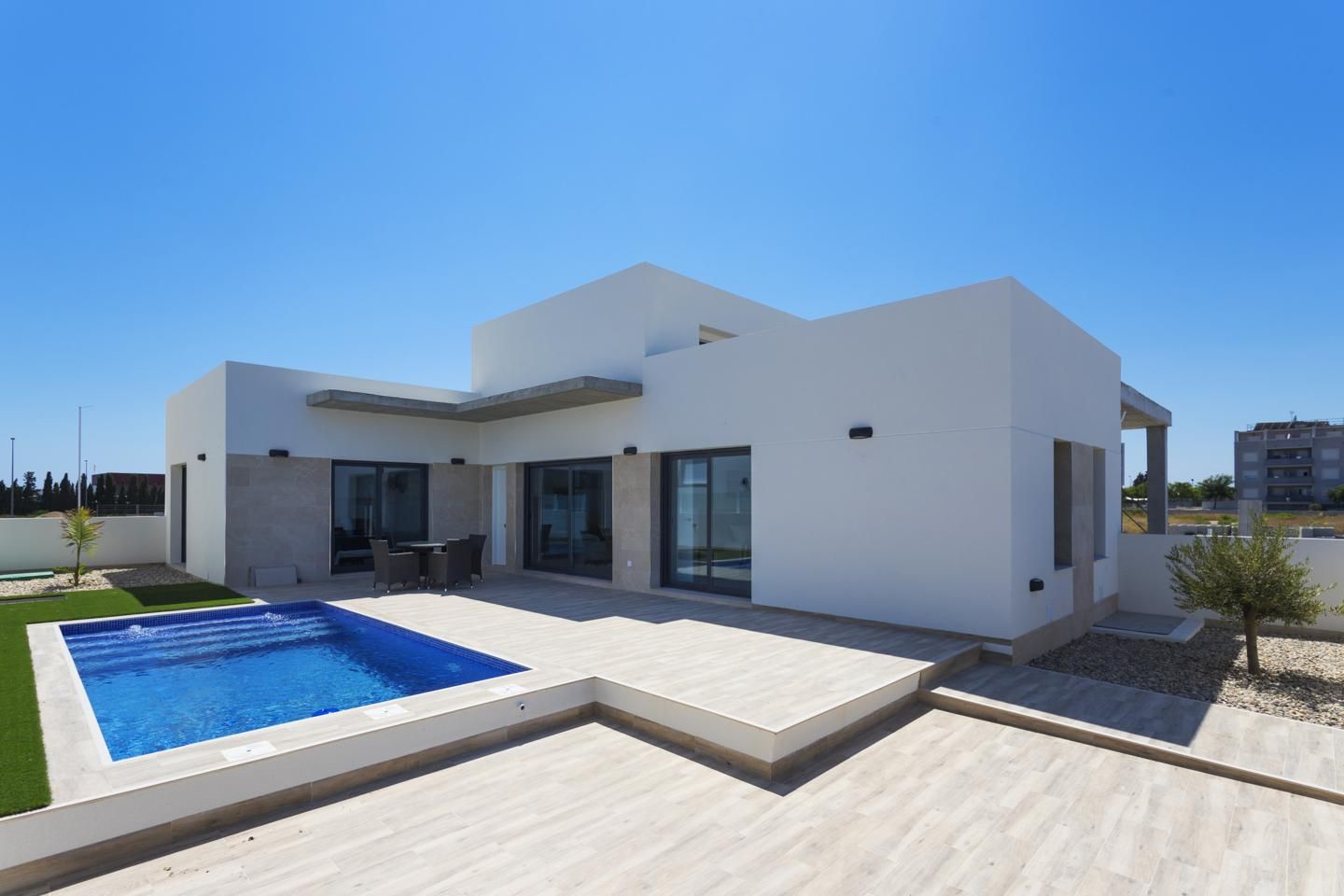 Modern white villa with a rectangular blue pool, glass doors, and a sunlit tiled patio.