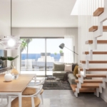 Bright modern living area with a wooden dining table, white chairs, and a gray sofa, lit by a pendant light and large glass doors to outdoors.