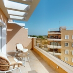Balcony with wicker chairs and glass table overlooking beige apartment buildings on a clear blue day.