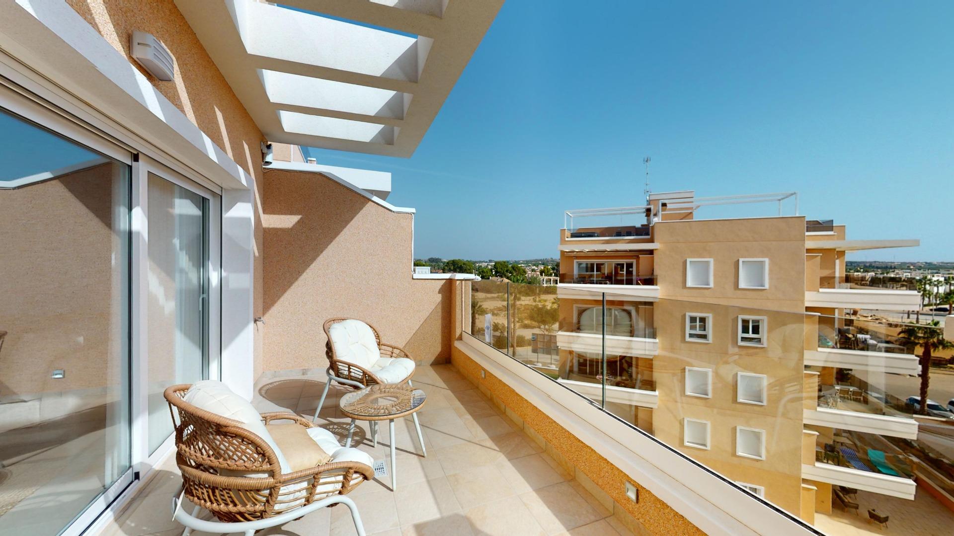 Balcony with wicker chairs and glass table overlooking beige apartment buildings on a clear blue day.