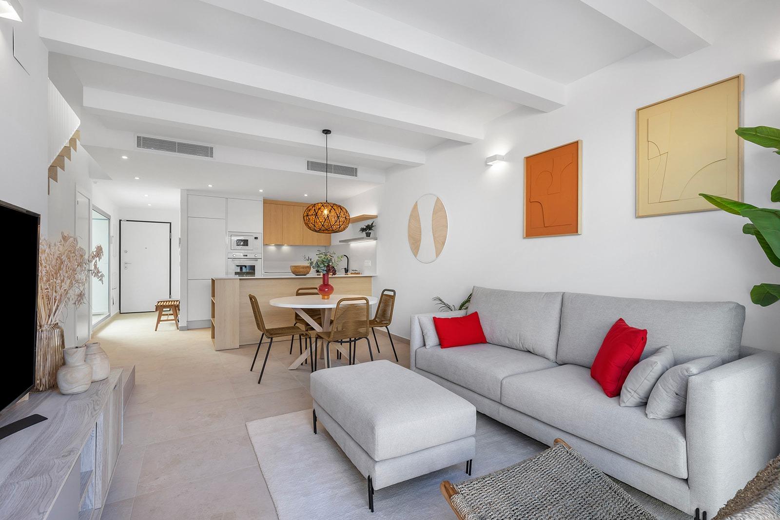 Open-plan living area with a light gray sofa and red cushions, a round dining table, and a woven pendant light near a white kitchen island; calm, modern decor in neutral tones with green plant accents.