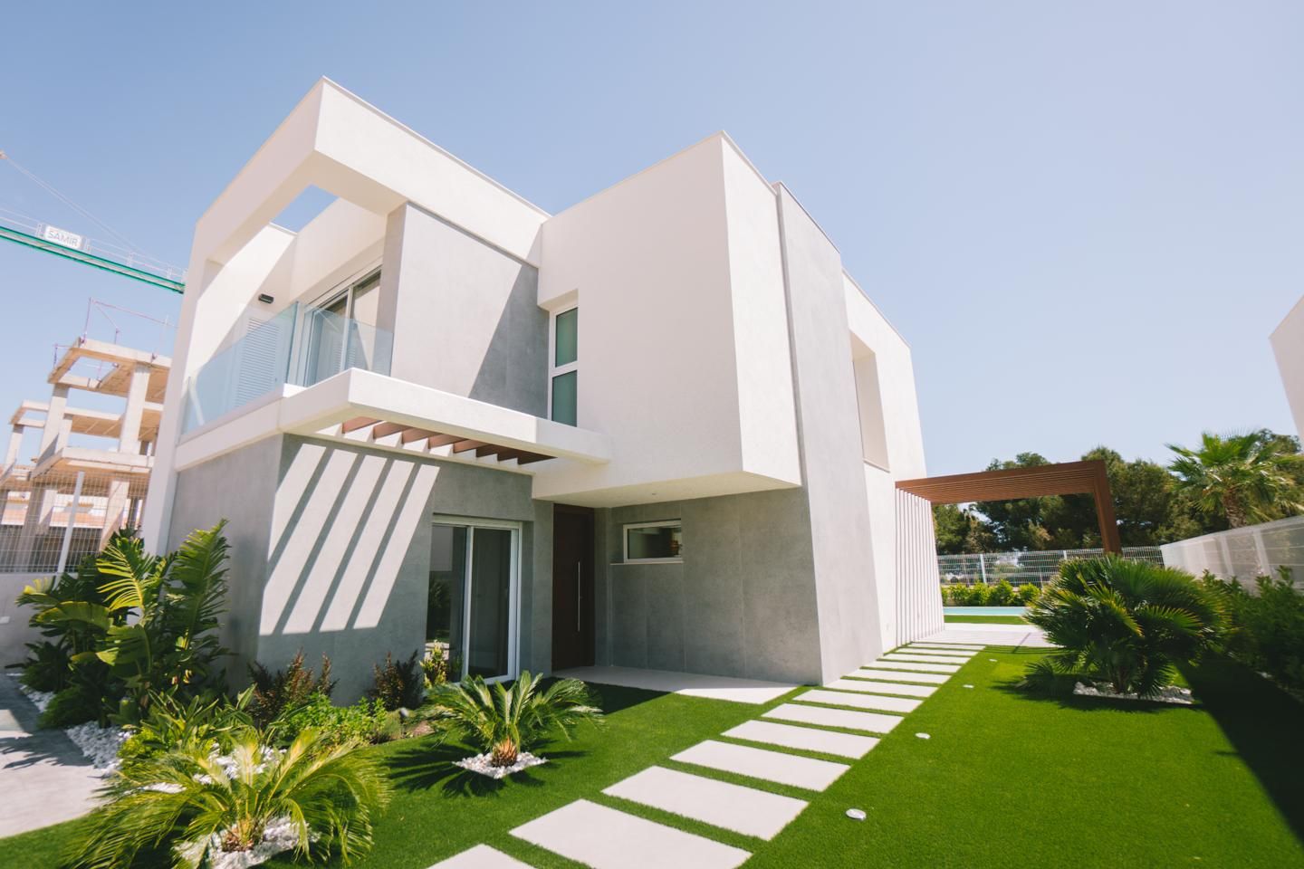 Modern white two-story house with glass balcony, grey accents, and a manicured lawn under a blue sky; stepping-stone path in foreground