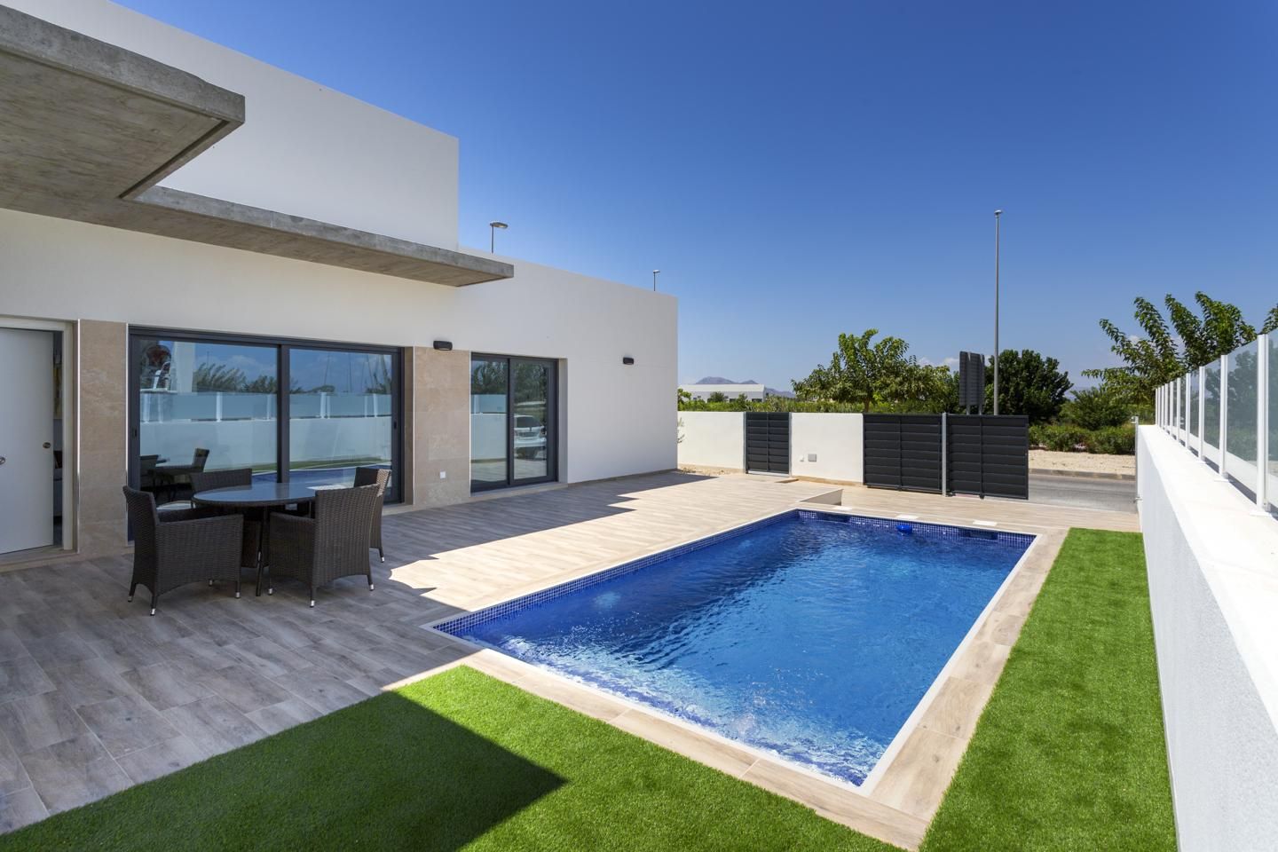 Modern backyard of a contemporary home with a rectangular blue pool, glass doors, and a wicker dining set on beige tile.