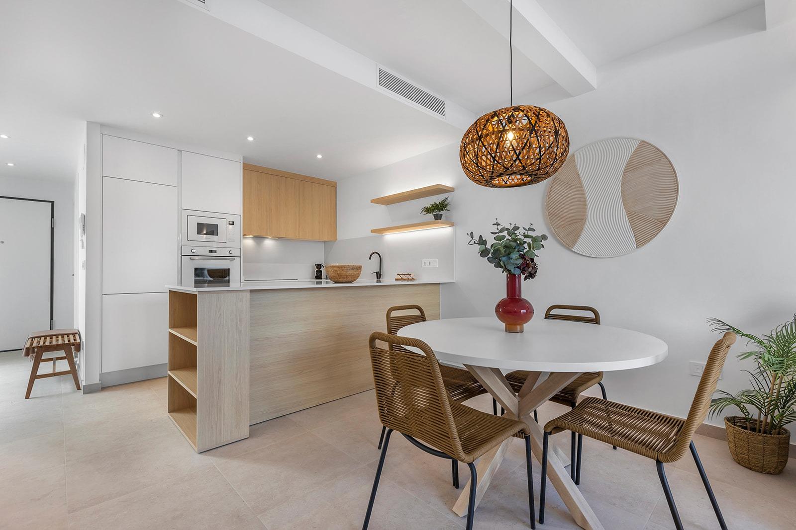 Open-plan kitchen with white cabinets and wood accents, featuring a round white dining table with four wicker chairs at the center