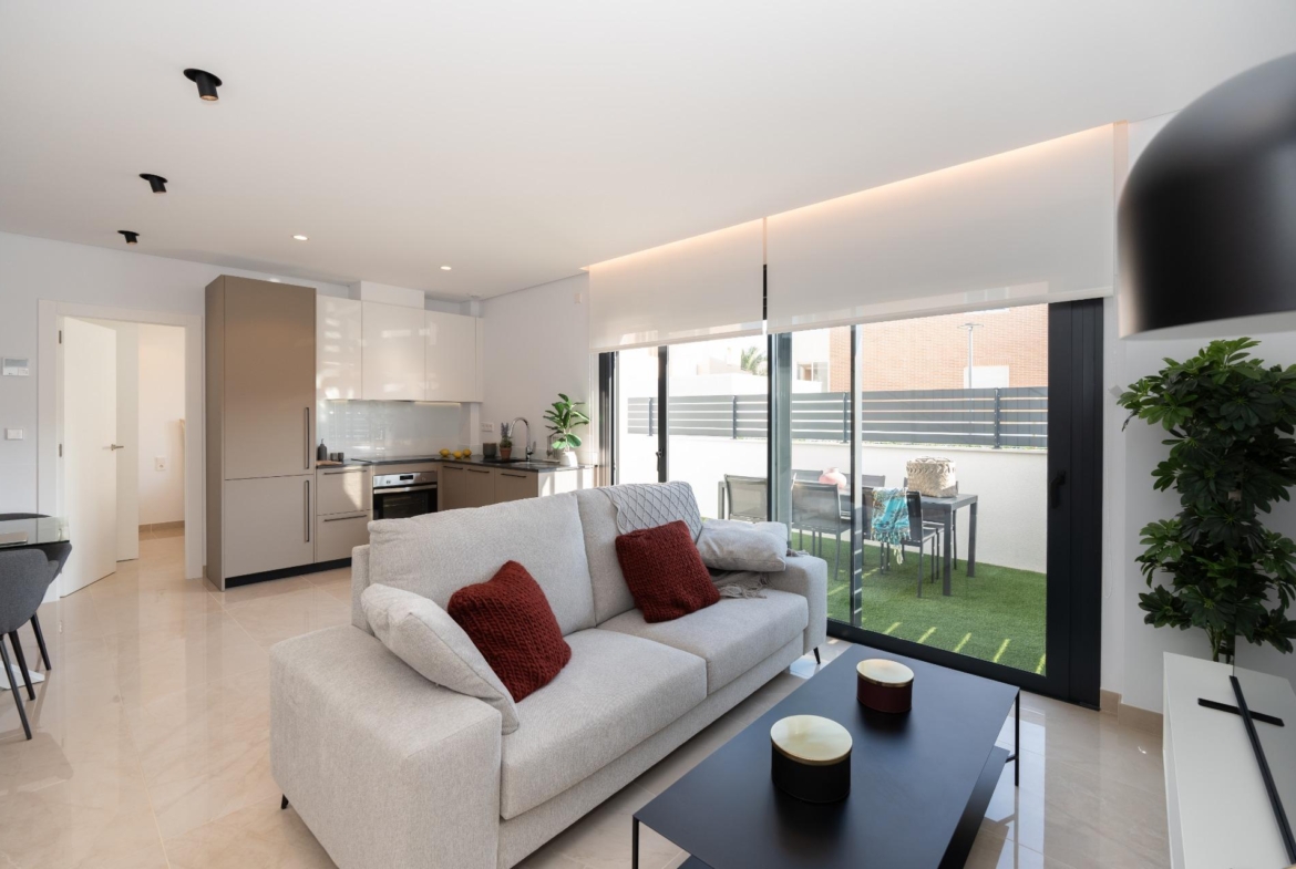 Open-plan living room with light gray sofa and red cushions facing a black coffee table, next to a modern kitchen and large glass doors to a patio garden outside.