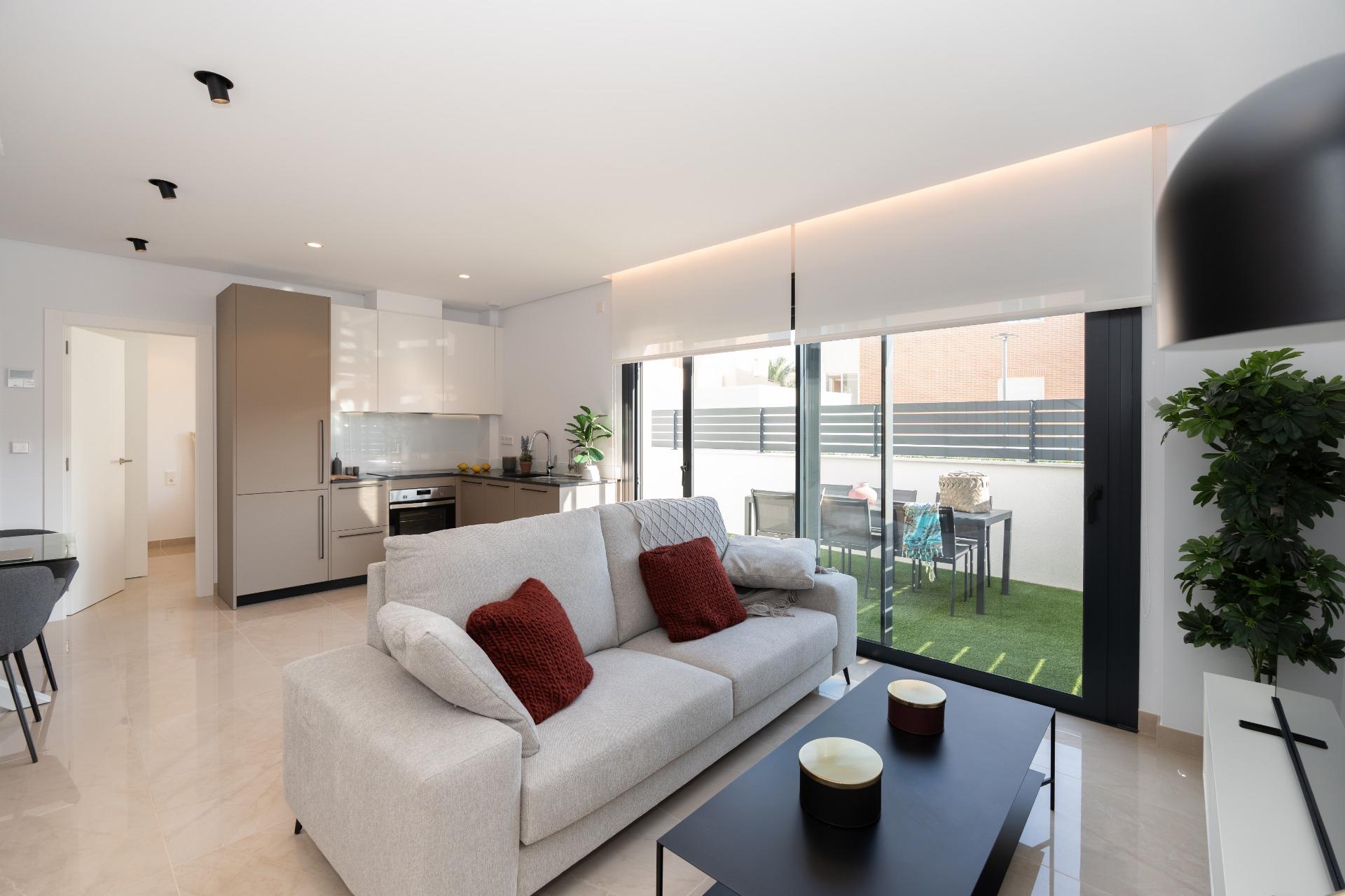 Open-plan living room with light gray sofa and red cushions facing a black coffee table, next to a modern kitchen and large glass doors to a patio garden outside.