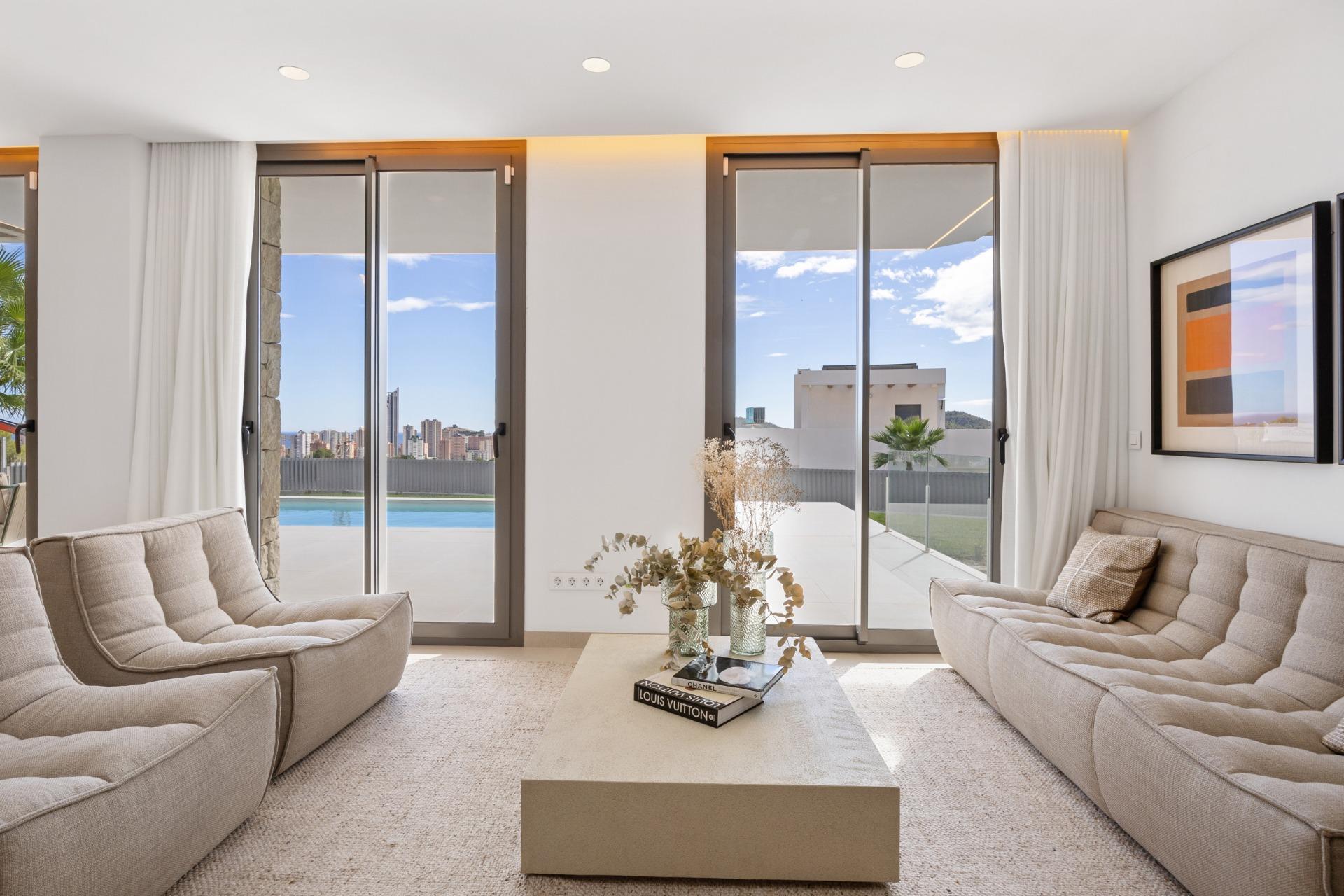 Bright modern living room with beige tufted sofas facing large sliding glass doors to a pool and city skyline outside.