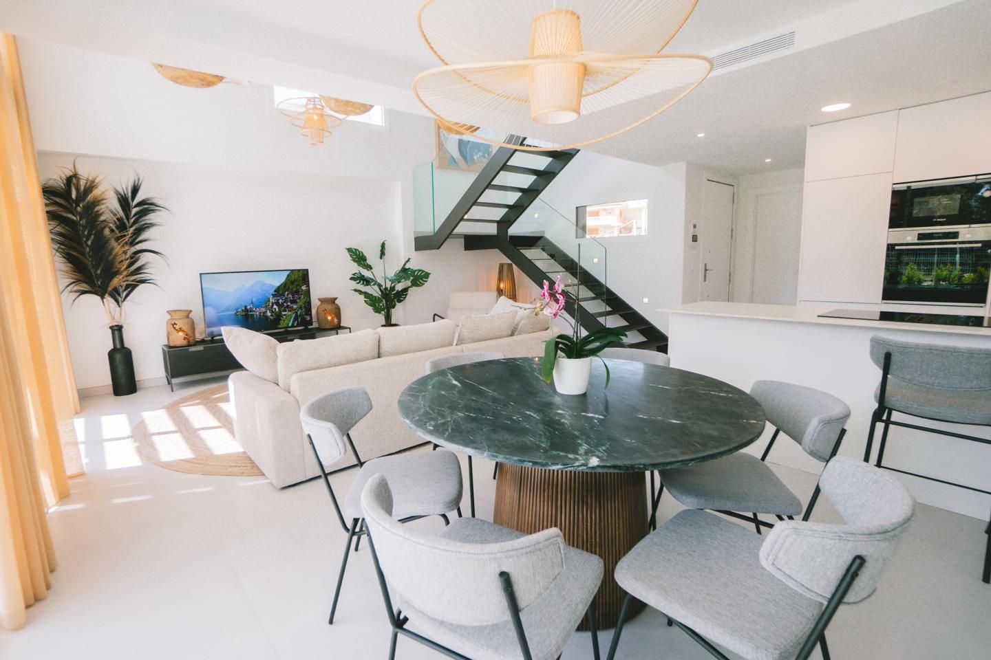 Open-concept living and dining area with a round green marble dining table, six gray upholstered chairs, a sofa, TV, and glass‑railed staircase.