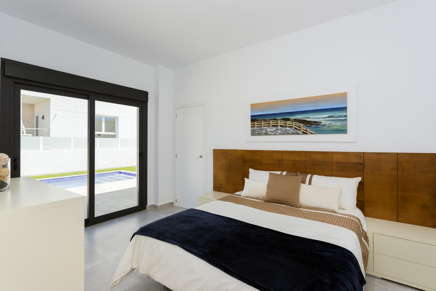 Modern bedroom with a large sliding glass door open to a pool area, white walls, and a wooden headboard over a neatly made bed.