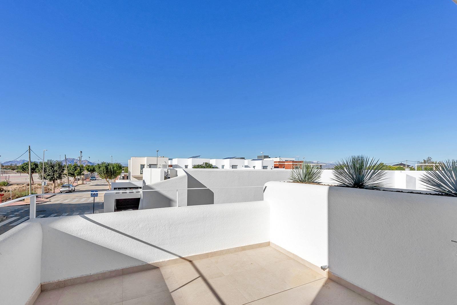 White cubic rooftops and walls rise across a sunny terrace, with desert plants and a bright blue sky overhead for a modern neighborhood view.