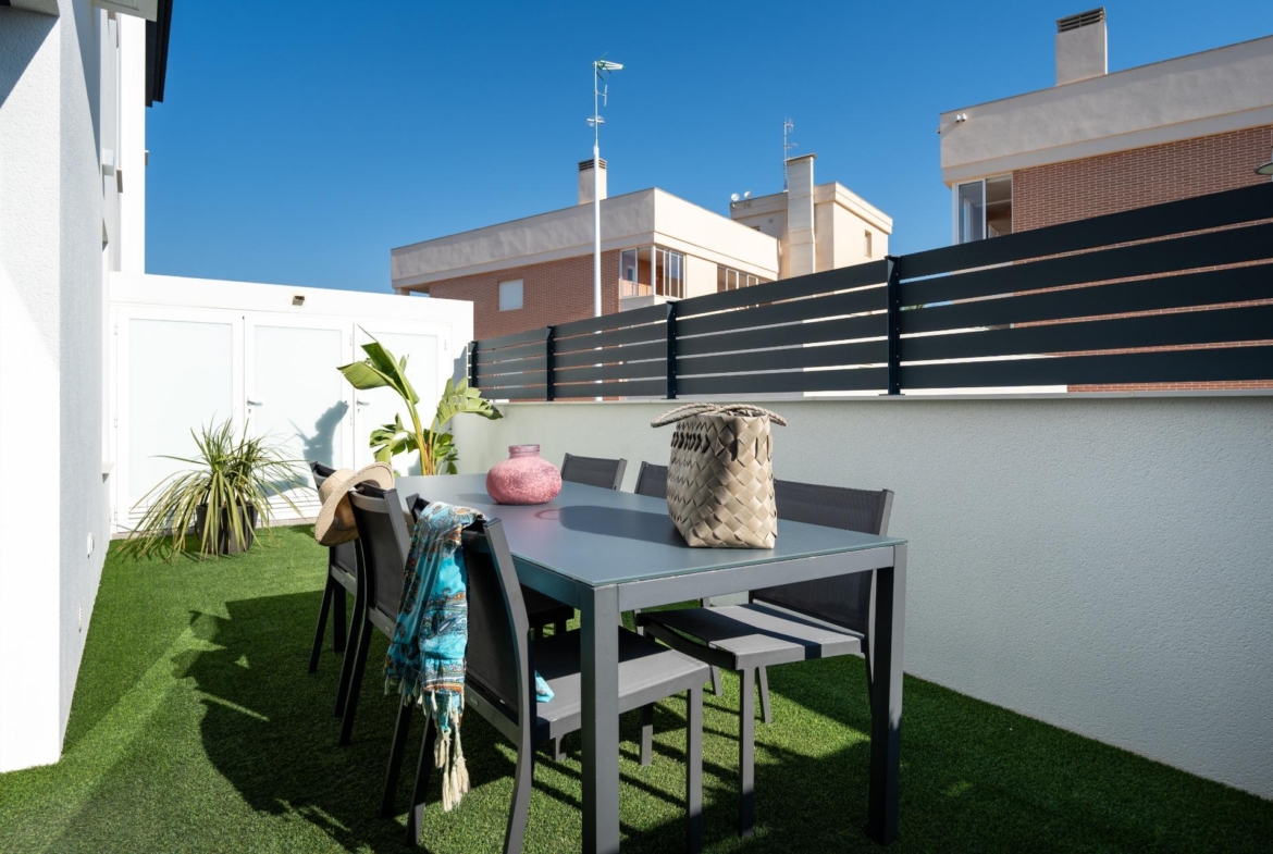 Rooftop patio with a gray dining table and chairs on green artificial turf, plants, and wicker decor against a dark slatted fence and neighboring buildings.