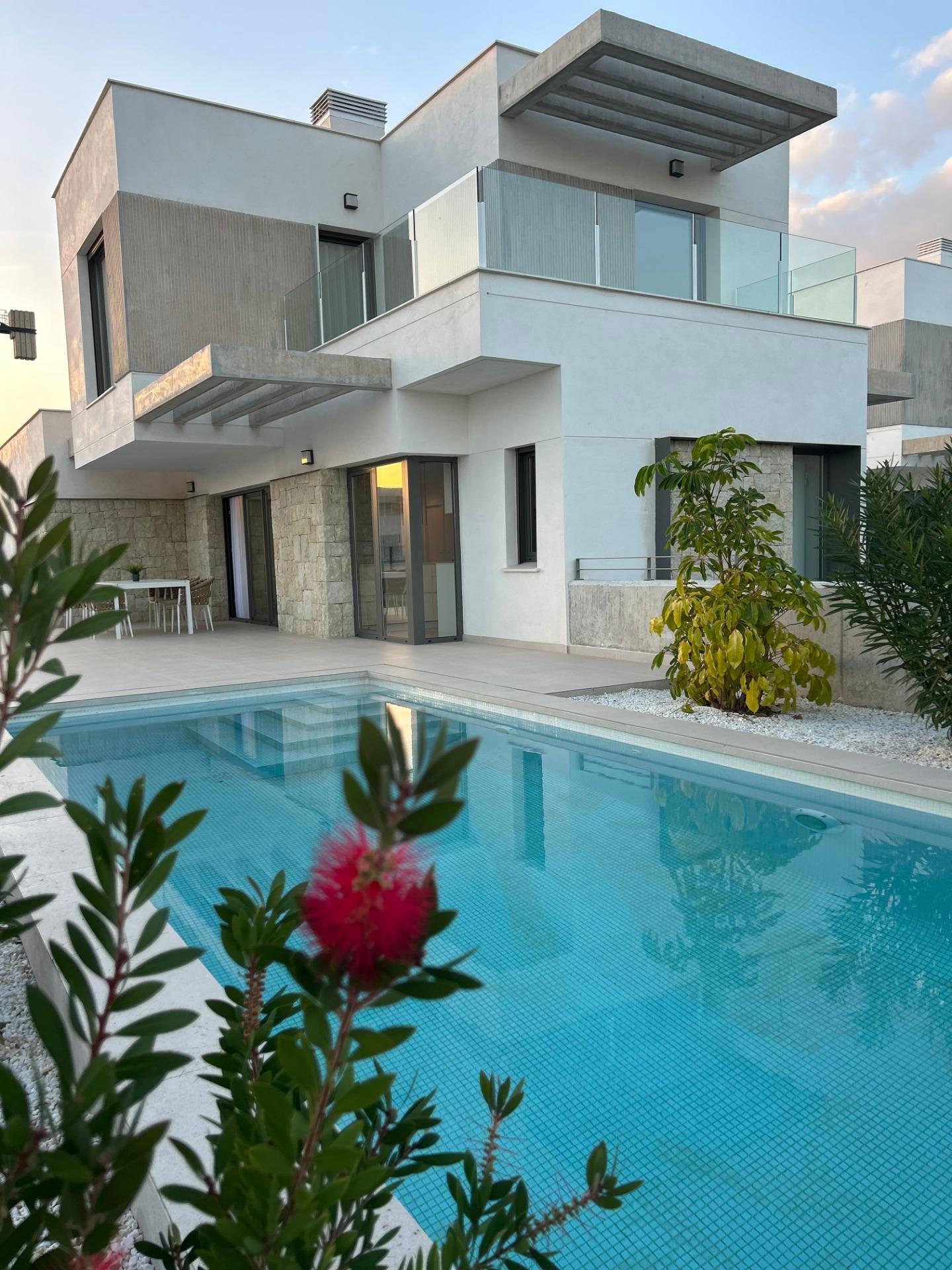 Modern white two-story house with glass railing, balcony area, and a rectangular pool in the foreground, with plants in the foreground.