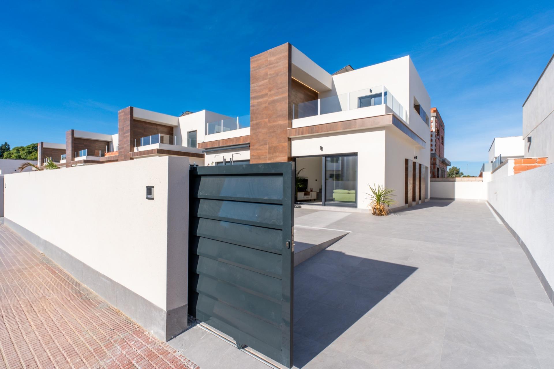 Modern white and brown townhouse complex with glass balconies seen from the entrance gate and paved driveway under a bright blue sky