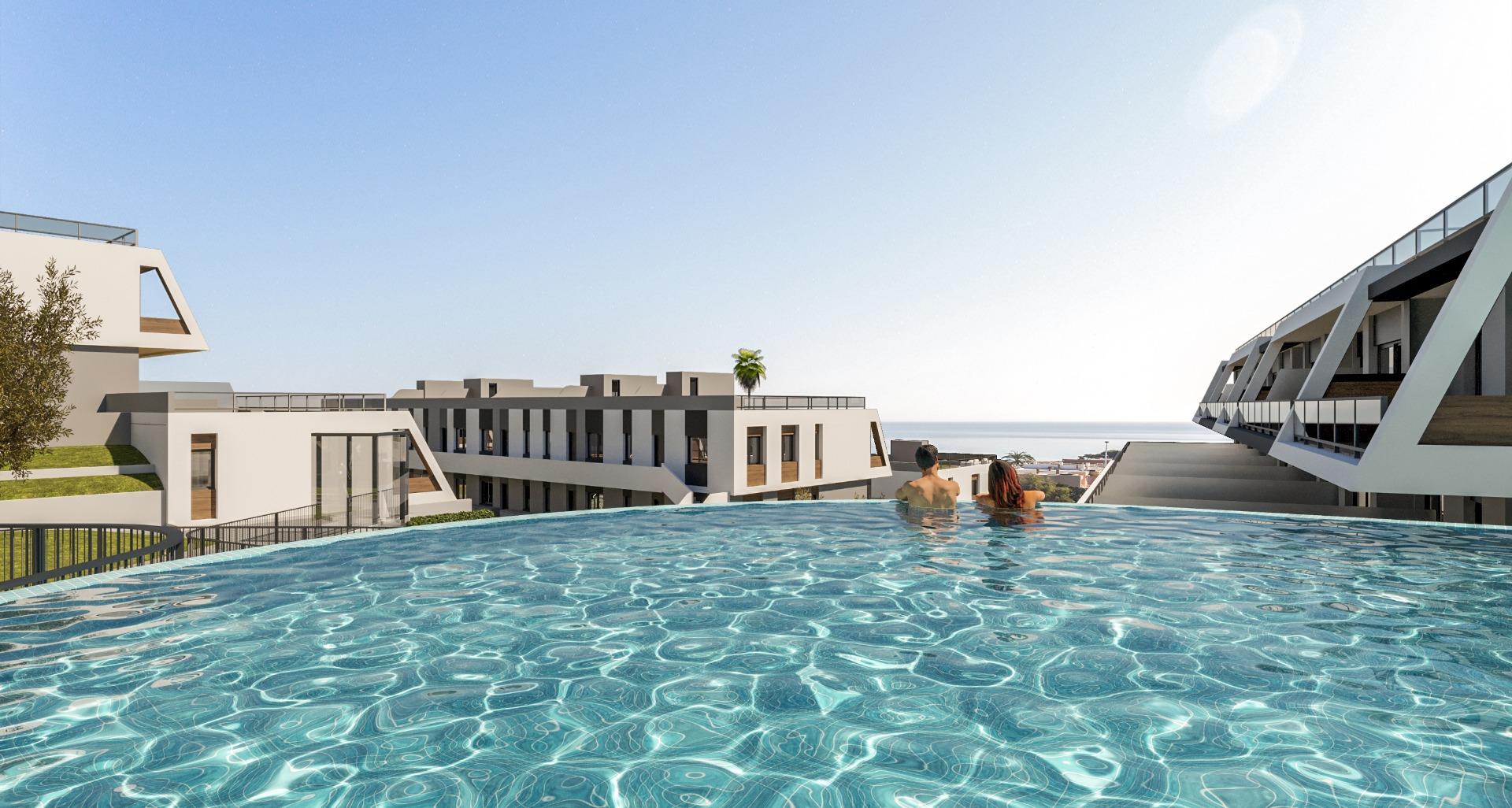 Infinity pool at a modern resort with two people relaxing in the foreground and white buildings against a clear blue sky and ocean in the distance.