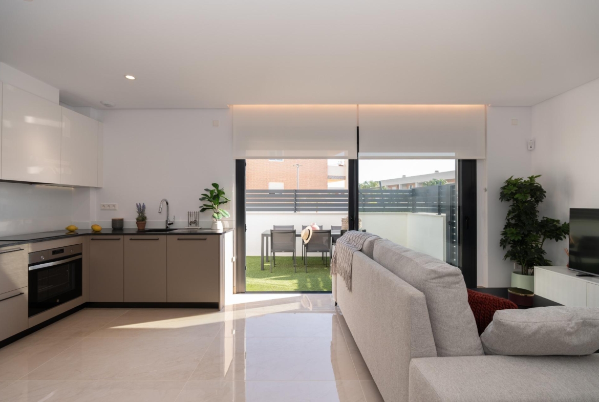 Open-plan kitchen and living room with white glossy cabinets, beige lower units, oven, sink, and a sliding glass door to the patio outdoor dining area at the back.