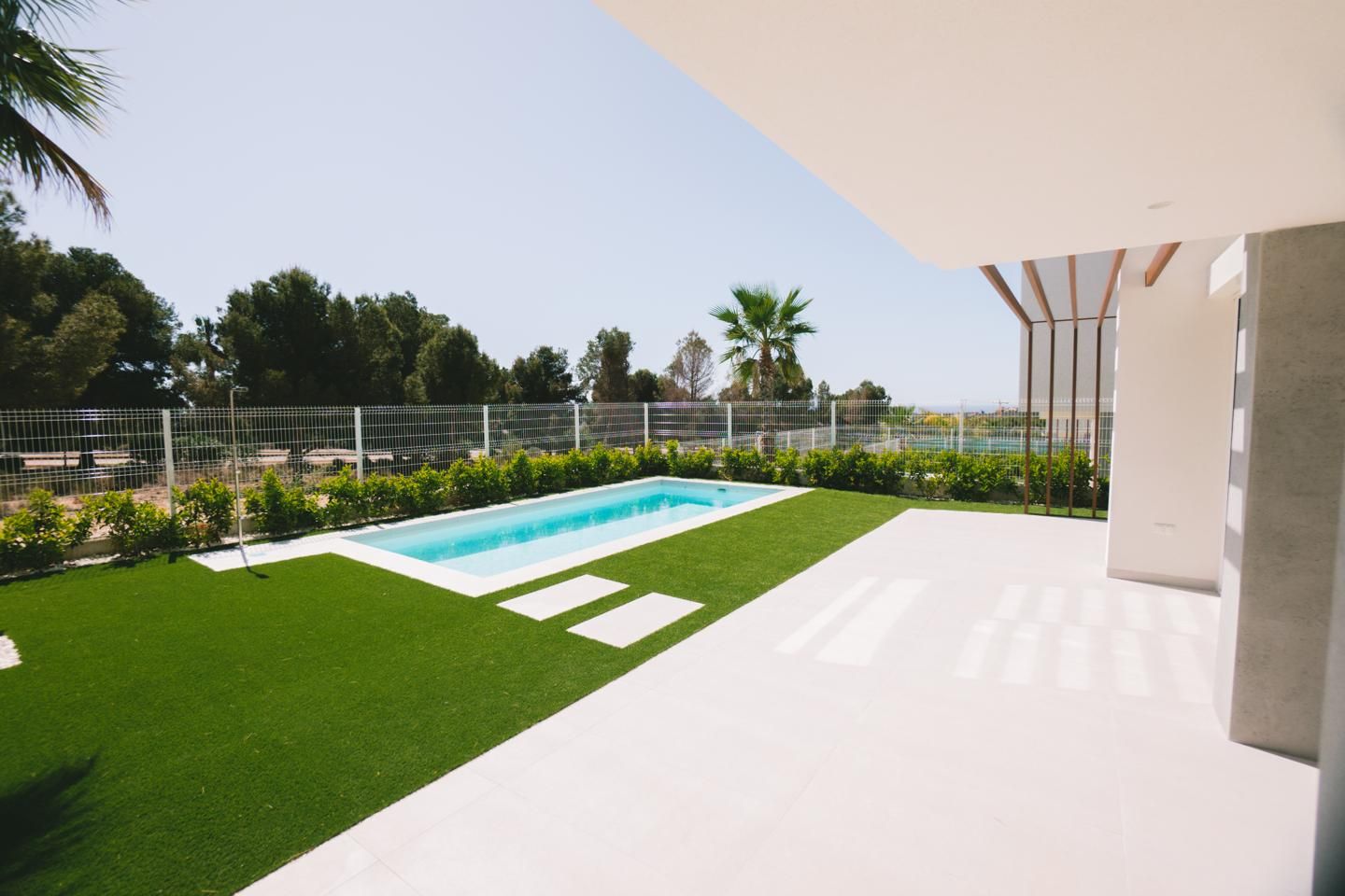 Backyard with rectangular pool, green lawn, white tiled patio, and a metal fence; palm trees in distance.