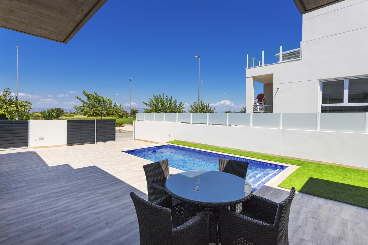 Modern backyard: rectangular blue pool, frosted glass panels, and a round glass table with wicker chairs on a gray tiled patio.