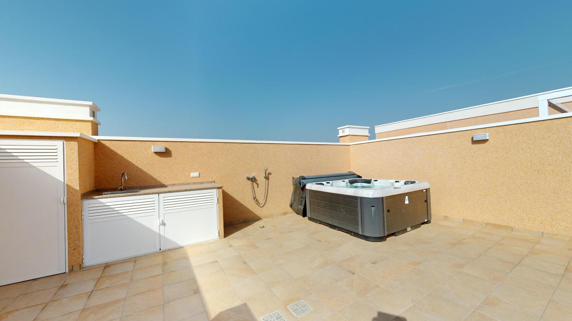 Rooftop terrace with a large hot tub on beige tiled floor and tan walls under a clear blue sky. A sink cabinet is on the left.