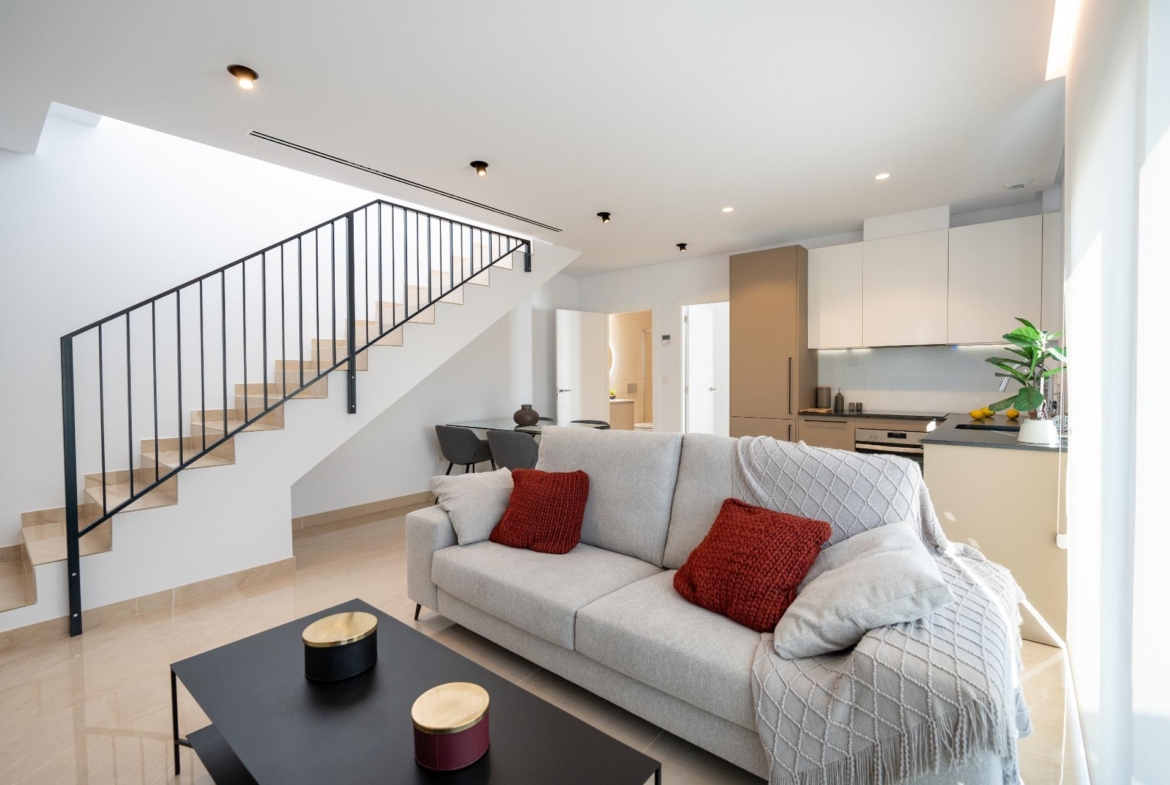 Open-plan living room with a beige sofa and red throw pillows, black coffee table, and a staircase along the left wall.",