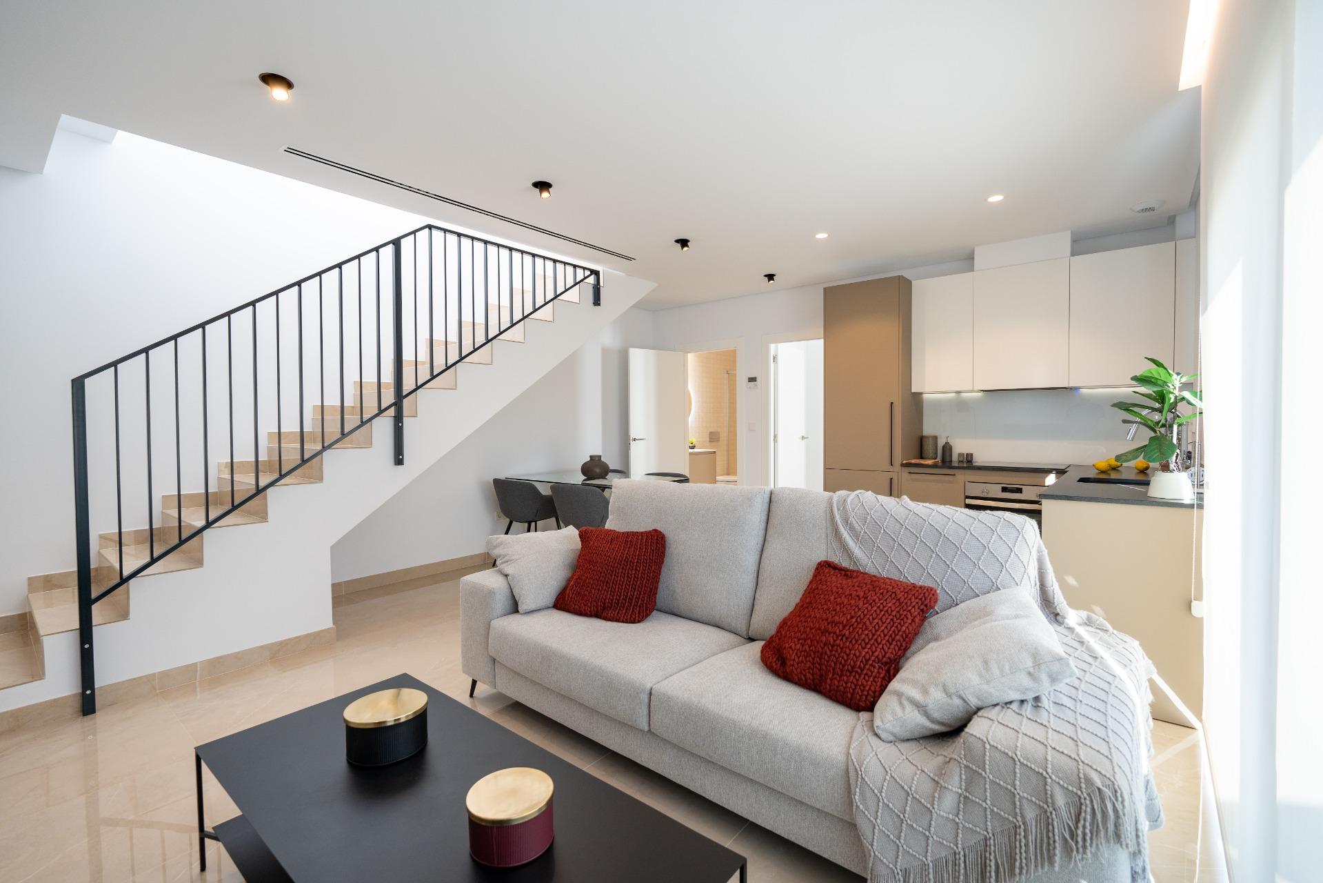 Open-plan living room with a beige sofa and red throw pillows, black coffee table, and a staircase along the left wall.",