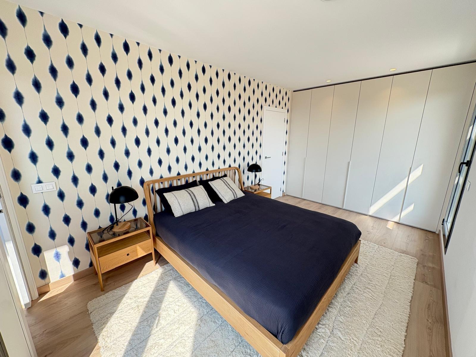 Bedroom with blue teardrop wallpaper, a wooden bed with navy bedding, two black bedside lamps, and a white rug.
