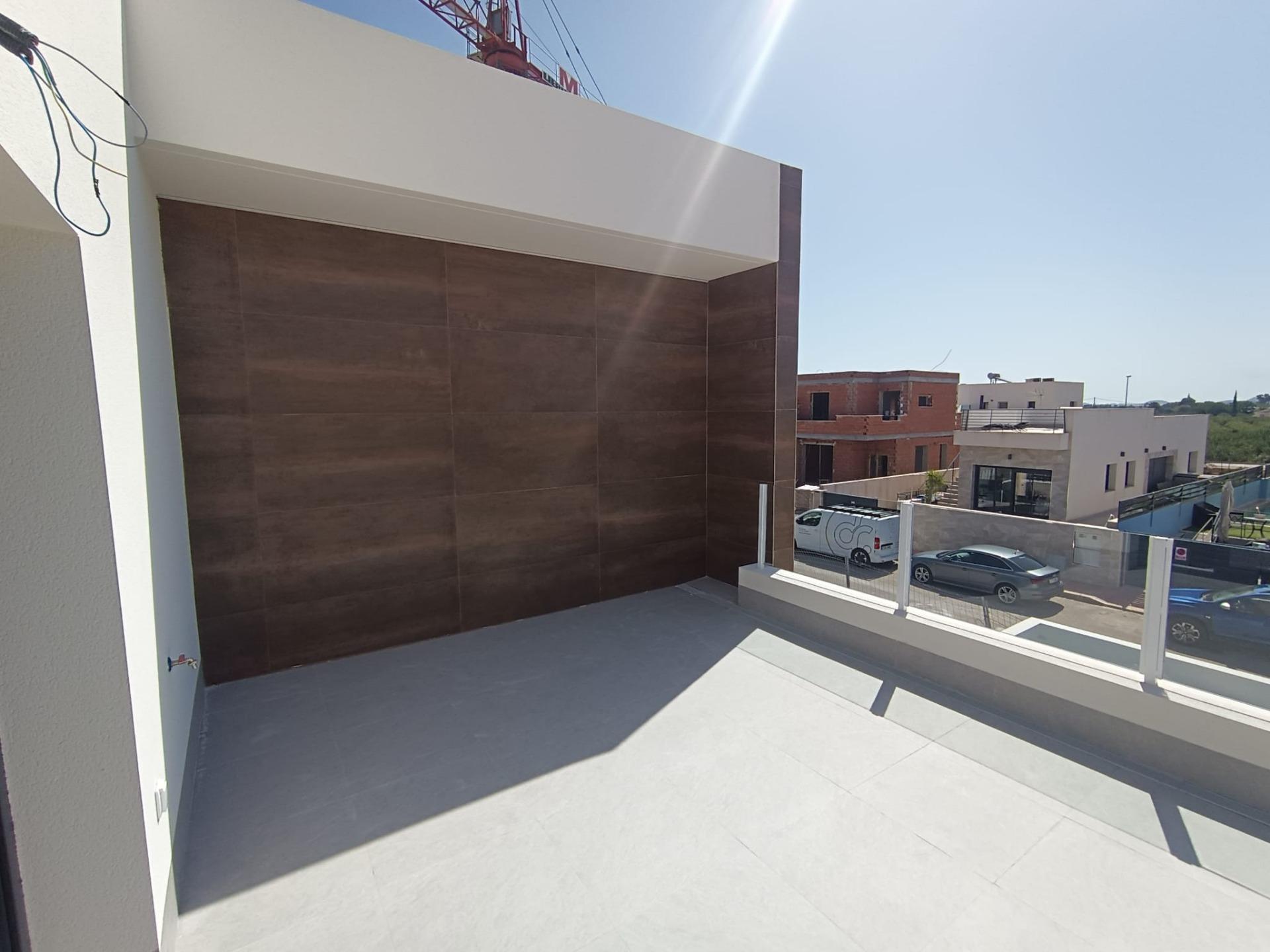 Modern rooftop terrace with gray flooring, brown tiled wall, and glass railing overlooking a suburban street with parked cars and houses in the distance.