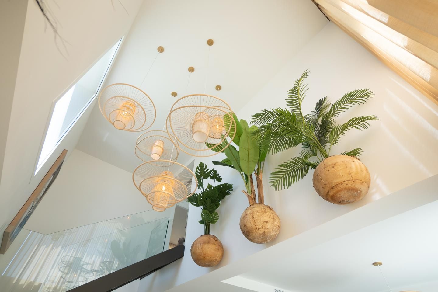 Bright modern lobby with three layered rattan circular pendant lights and large potted palms in coconut planters on a white wall.