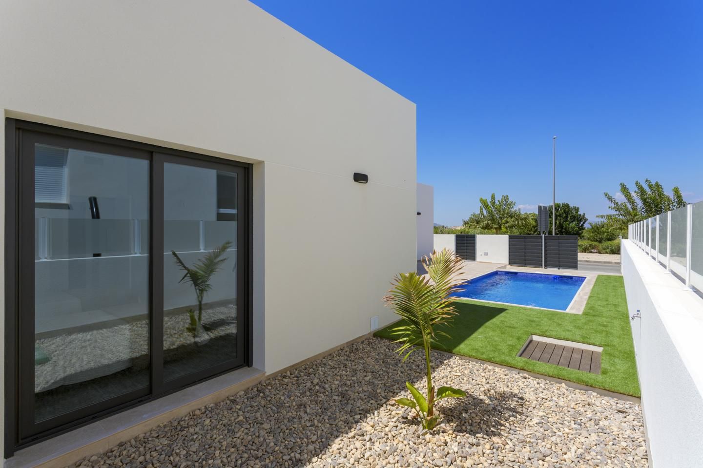 Modern backyard with sliding glass doors, a rectangular blue pool, and a gravel path beside artificial turf under a clear blue sky