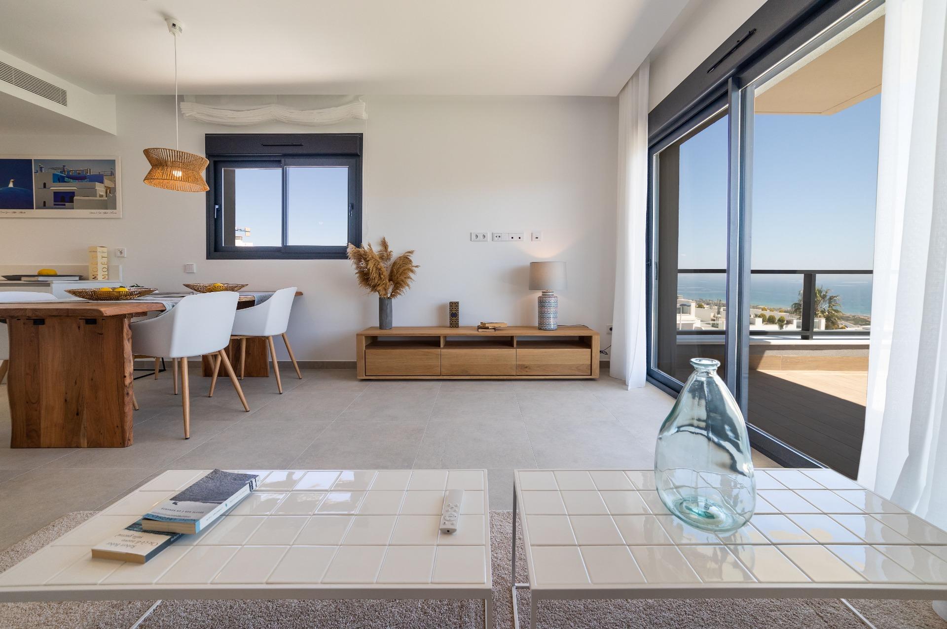 Bright open-plan living and dining area with white tiled coffee tables, a wooden dining table with white chairs, and a balcony door revealing the sea view.