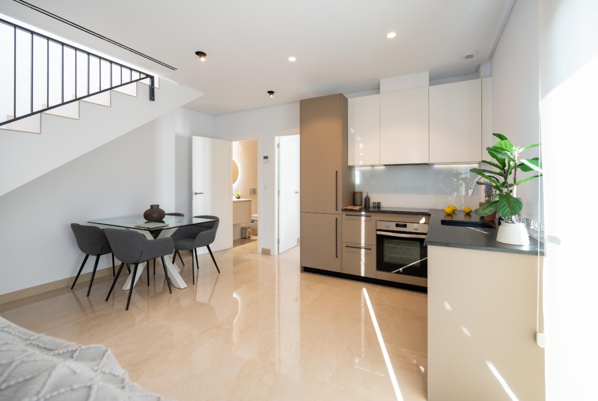 Open-plan kitchen and dining area with beige cabinetry, stainless appliances, glass dining table, and a staircase along the left wall under bright lighting.