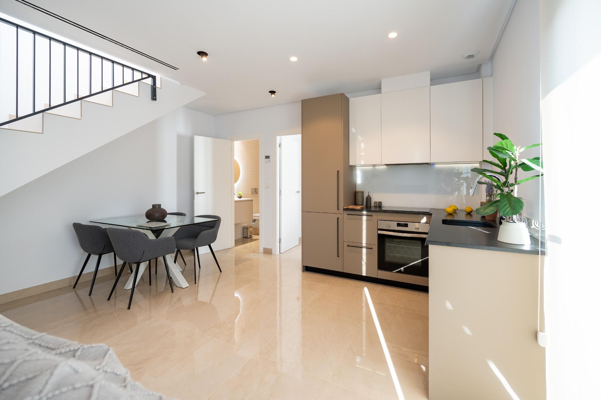 Open-plan kitchen and dining area with beige cabinetry, stainless appliances, glass dining table, and a staircase along the left wall under bright lighting.