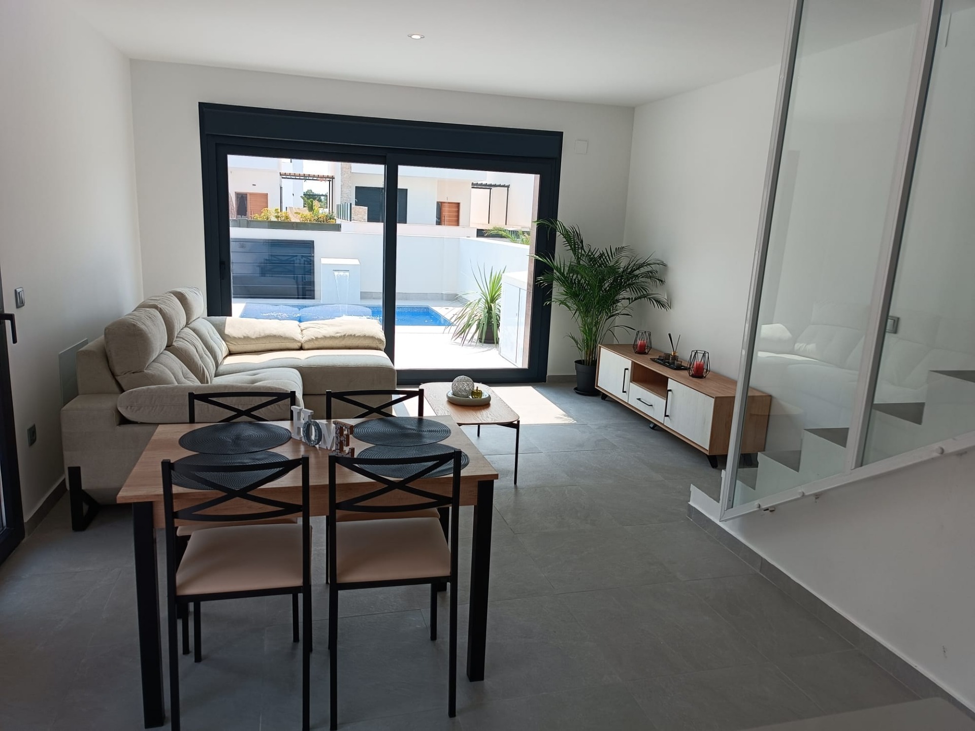 Open-plan living and dining area with a beige sectional sofa, wooden dining table for four, and sliding glass doors to a pool outside.