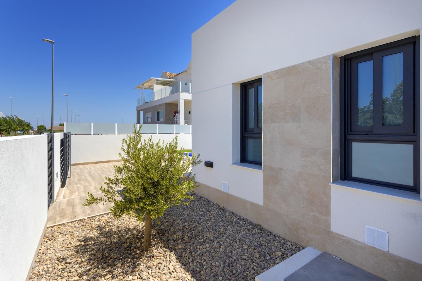 Modern white house exterior with beige stone accents, two dark-framed windows, and a small olive tree in a gravel yard.
