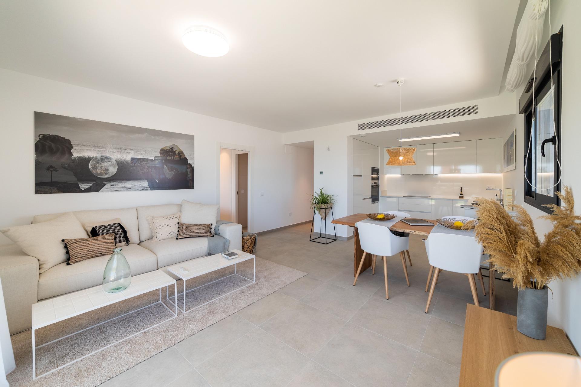Bright open-plan living area with a beige sectional, white coffee tables, wall art, and a doorway to the kitchen beyond, plus a dining table nearby.