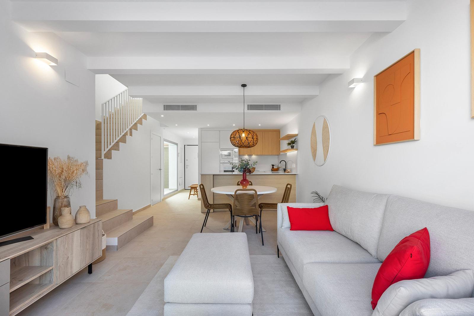 Open-plan living room with a gray sectional, red cushions, and a round white dining table, kitchen in the background.