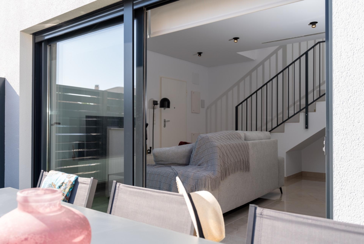 Living room seen through sliding glass doors: gray sofa, black metal staircase railing, white walls, and ceiling lights outside.