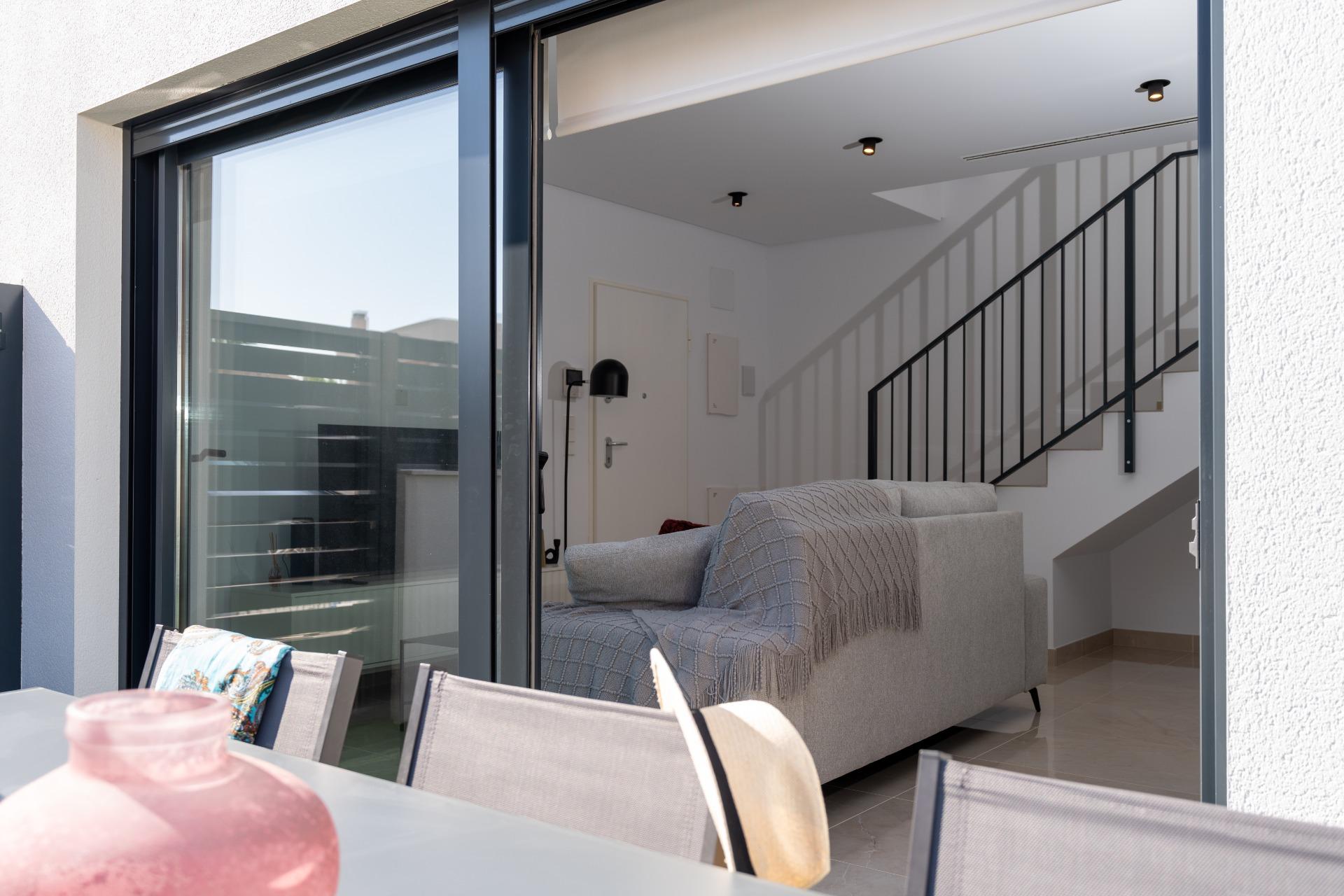 Living room seen through sliding glass doors: gray sofa, black metal staircase railing, white walls, and ceiling lights outside.