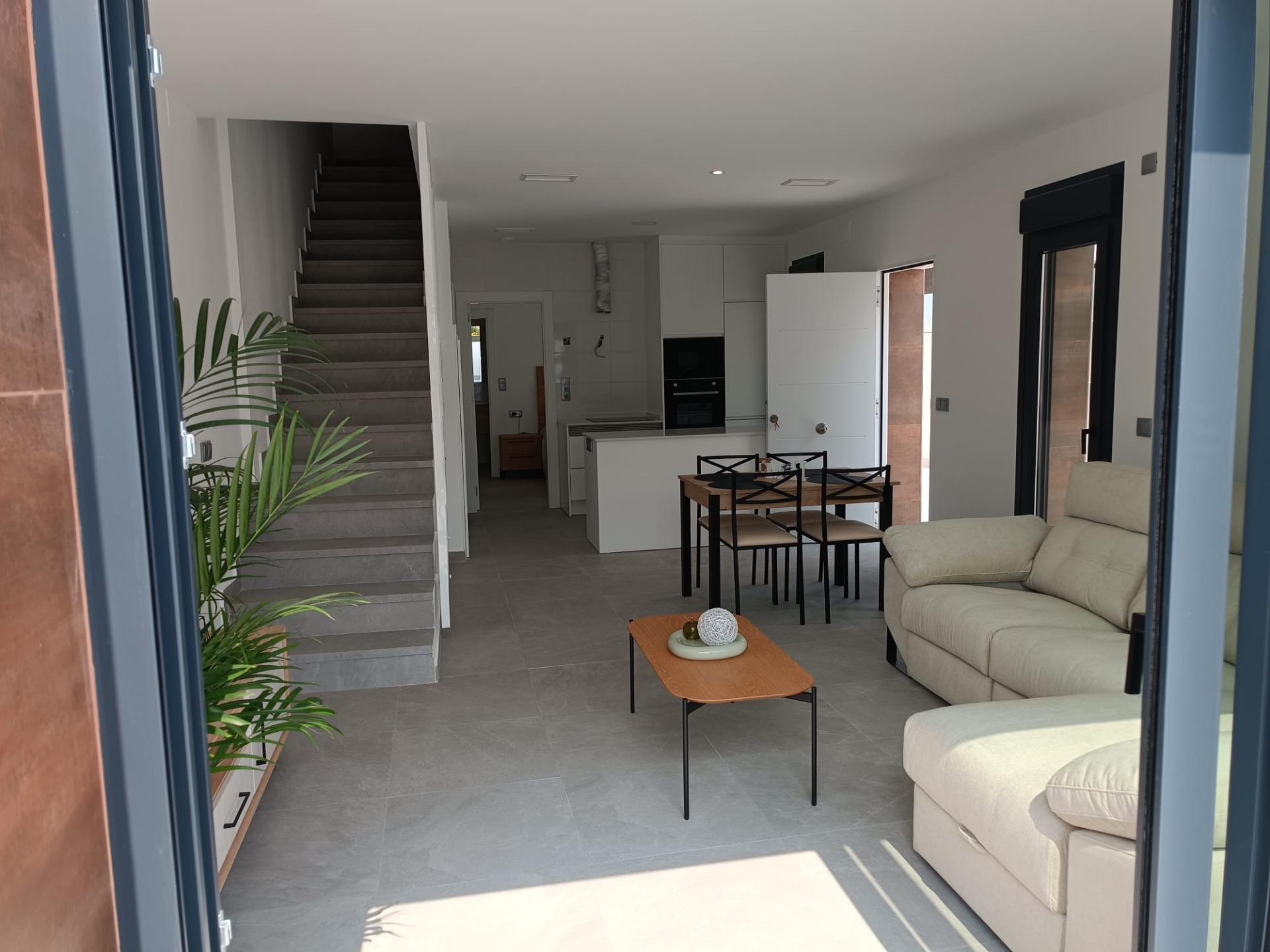Open-plan living area with a beige sofa, wooden dining table, and a white kitchen in the background; stairs on the left side.