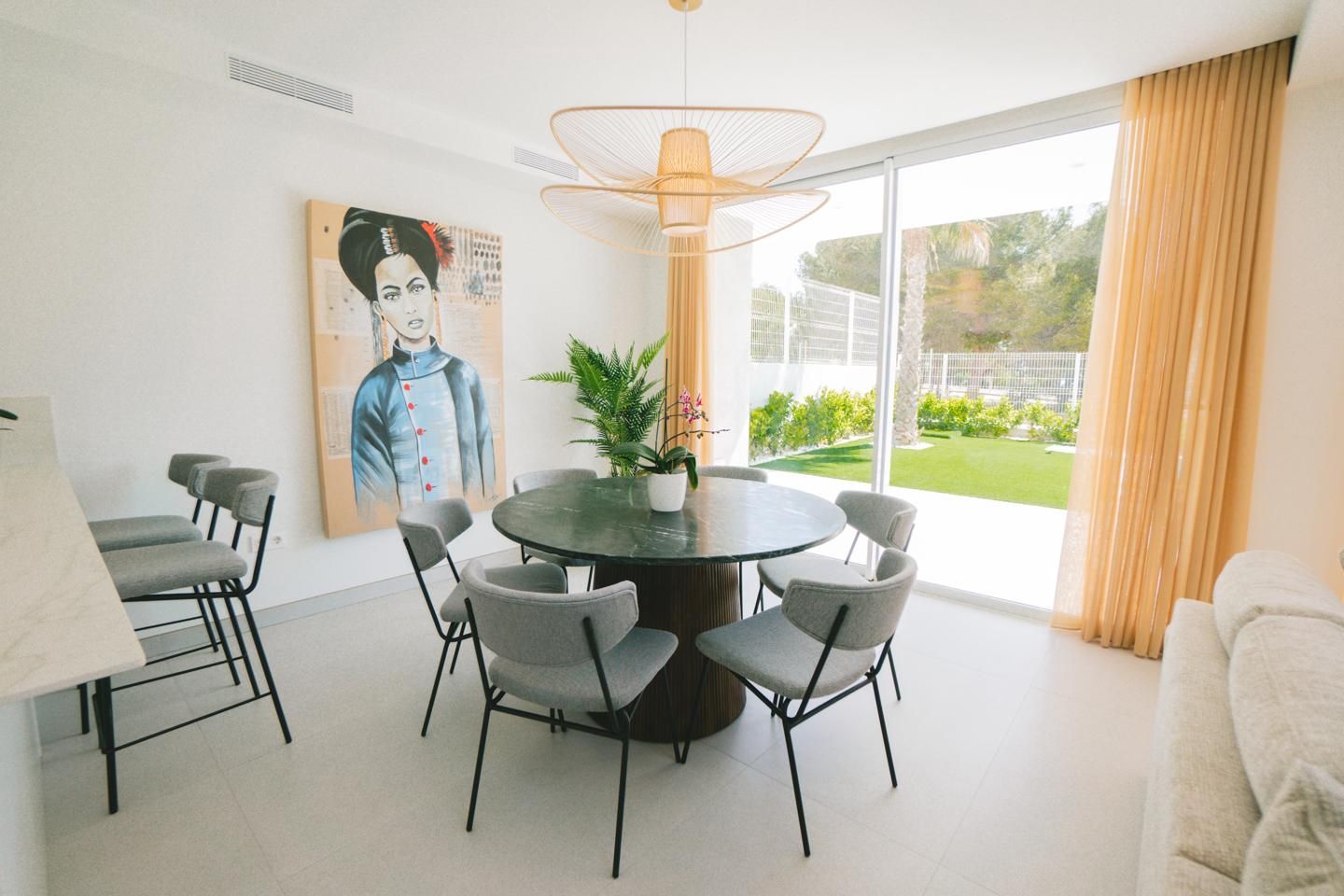 Bright dining area with a round dark marble table surrounded by grey upholstered chairs, a large wall portrait, and floor-to-ceiling windows with peach curtains opening to a green garden.